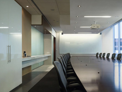 An empty conference room with long table and chairs on each side. A blank whiteboard is on the far wall, and projector attached to the ceiling.