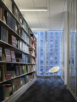 Library with tall shelves in high-rise building, with empty white chair. Floor to ceiling windows show other tall buildings outside.