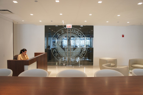 Woman seated behind reception desk in an office building with large glass doors behind her.