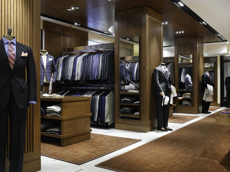 Men's formalwear store with custom woodwork on walls. Racks of clothing are inset into another room that also has a wooden stand with folded shirts. Suited mannequins stand along the wall.t