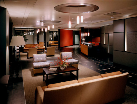 Modern office receiption area with low seating couches and armchairs, floor lamps and hanging lighting. A brown coffee table has a book and red flowers on it.