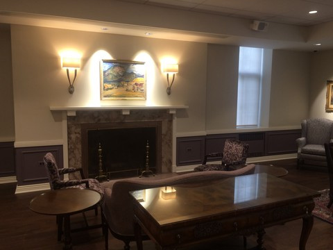 Custom woodwork in a hearth room. Landscape are is displayed over a fireplace with lit sconce lamps on either side. A couch and two chairs are in front of the fireplace.